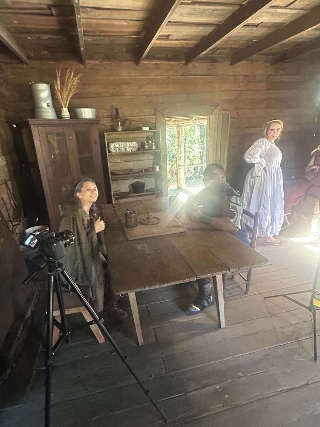 Costumed cast gathered around a table inside the cabin, camera on a tripod in the foreground