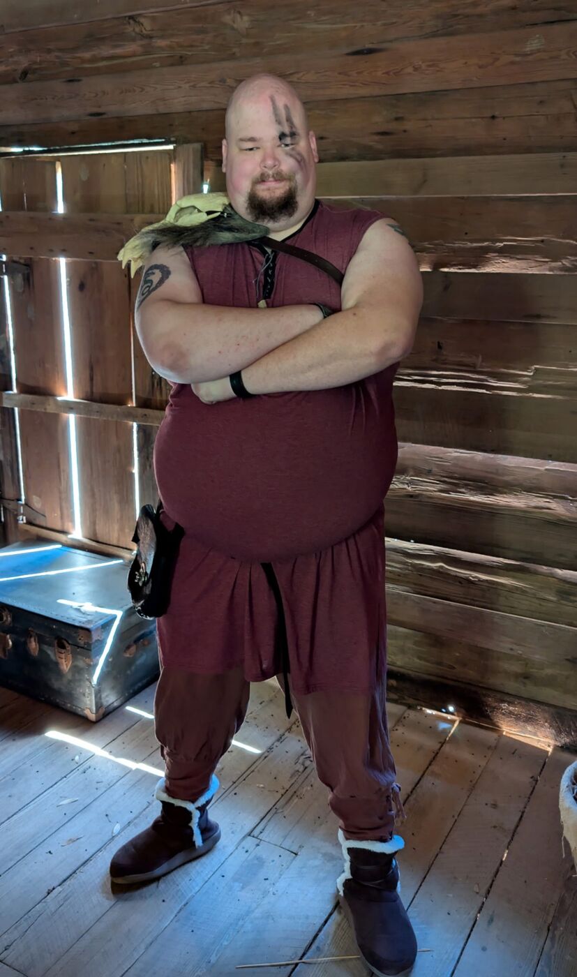 A costumed warrior standing with arms crossed in the timber cabin, sunlight through the walls